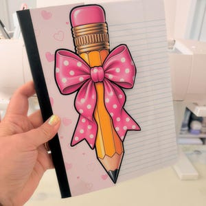 May include: A notebook with a black spine and a cover featuring a light pink background with small hearts. The cover displays a large illustration of a yellow pencil with a pink eraser and a bright pink bow with white polka dots on top.