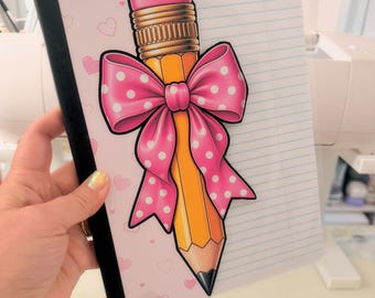 May include: A notebook with a black spine and a cover featuring a light pink background with small hearts. The cover displays a large illustration of a yellow pencil with a pink eraser and a bright pink bow with white polka dots on top.