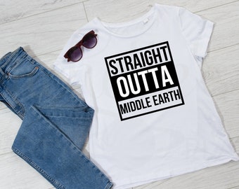 May include: A white t-shirt with the text "STRAIGHT OUTTA MIDDLE EARTH" in a black box, paired with blue jeans and sunglasses. The sunglasses are dark brown. The items are arranged on a light wood-grain surface.
