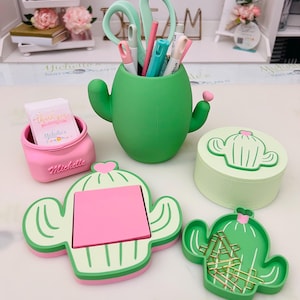 May include: A collection of desk accessories in a cactus theme. Includes a green cactus-shaped pen holder with scissors and pens, a pink container with "Thank You" cards, a cactus-shaped notepad, and a cactus-shaped paperclip holder.