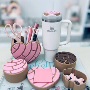 Op de afbeelding: Een witte Stanley-beker met een roze deksel en rietje, naast een hartvormige bureau-organizer en andere accessoires. De organizer en andere containers hebben een concha-broodontwerp in roze en bruin. Pennen, scharen en plaknotities zijn ook zichtbaar.
