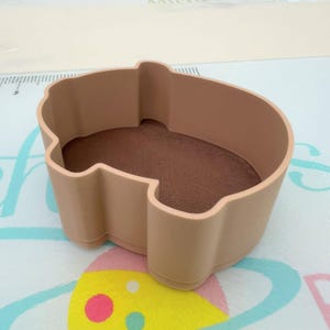May include: A tan-colored, pig-shaped cookie cutter. The interior is a darker brown color. The cutter is made of a sturdy material and is designed for baking. The cutter is sitting on a patterned surface.