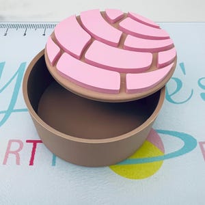 May include: A brown container with a pink lid. The lid is round and has a design resembling a concha pastry. The container is open, revealing its interior. The background is a patterned surface with text.