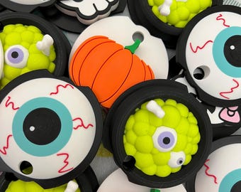 May include: A collection of Halloween-themed bottle stoppers. The stoppers feature designs such as a green, textured monster with a single eye, a white skull with a pink bow, a bright orange pumpkin, and a white eyeball with a blue iris.