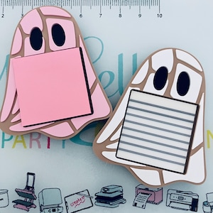 May include: Two ghost-shaped memo holders. One is pink with a pink sticky note, the other is white with a lined notepad. Both have black eyes and a tan outline. The memo holders are on a light blue surface with office supply illustrations.