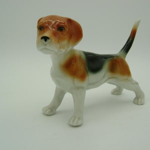 Puede incluir: Figura de cerámica de un perro beagle en posición de pie. El perro tiene un cuerpo blanco con marcas marrones y negras. El beagle tiene la cabeza y las orejas marrones, una nariz negra y una cola con punta negra.