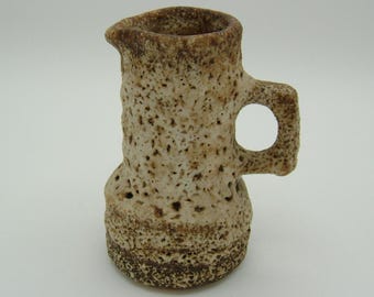 May include: A small, white ceramic pitcher with a textured surface and a brown handle. The pitcher has a round base and a slightly flared top.