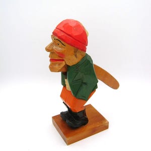 Op de afbeelding: Houten notenkrakerfiguur die een man afbeeldt met een rode hoed, groene jas en oranje broek. De figuur staat op een kleine houten basis en heeft zwarte laarzen. De mond van de notenkraker staat open.