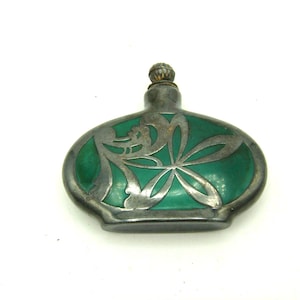 Op de afbeelding: Een ovaalvormig parfumflesje met een diepgroene, doorschijnende behuizing, omhuld door een zilverkleurige metalen overlay met een bloemrijk uitgesneden ontwerp. Het flesje heeft een kleine, getextureerde, ronde metalen dop en een platte bodem.