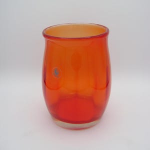 Op de afbeelding: Een helderoranje glazen vaas met een afgeronde vorm en een licht uitlopende rand. Het glas loopt van dieprood aan de basis over in een lichtere oranje kleur aan de bovenkant. Een kleine, ronde blauwe sticker is zichtbaar aan de zijkant.
