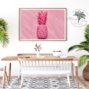 May include: A pink pineapple on a pink background with a subtle diagonal pattern. The pineapple is in a framed print with a white mat and a wood frame. The print is hanging on a white wall above a wooden table with a white chair.