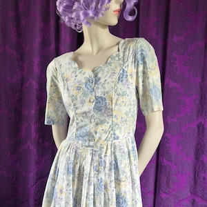 May include: A vintage floral print dress with a scalloped neckline and short sleeves. The dress has a button-down bodice and a pleated skirt. The fabric is white with blue, yellow, and purple floral patterns. The dress is displayed on a mannequin.
