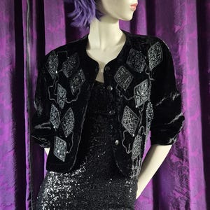 Könnte beinhalten: Ein schwarzes Pailletten-Minikleid mit einem schwarzen Samt-Bolero. Die Jacke hat rautenförmige Verzierungen. Die Mannequin hat lila Haare und steht vor einem lila Vorhang.