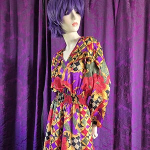 Puede incluir: Vestido vintage de manga larga con cuello en V y cintura elástica. El vestido presenta un vibrante estampado de formas geométricas y diseños florales rojos, amarillos, morados y negros. El vestido se exhibe en un maniquí.