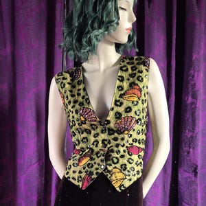 May include: A sleeveless vest with a V-neck, featuring a light green background with a leopard print pattern. The vest is adorned with seashell designs in shades of orange, pink, and purple. It has a button closure and is worn over a dark brown garment.