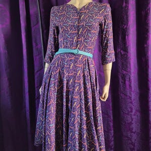 May include: A vintage purple dress with a colorful abstract pattern. The dress features a button-down front, three-quarter sleeves, and a flared skirt. A light blue belt cinches the waist. The dress is displayed on a mannequin.