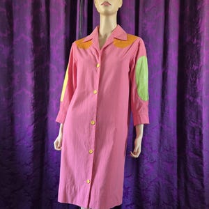 Puede incluir: Un vestido rosa con botones, cuello y mangas cortas. El vestido presenta botones dorados y parches de tela naranja y verde en los hombros y las mangas. El vestido se exhibe en un maniquí.