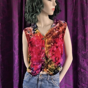 May include: A sleeveless vest featuring a vibrant, multi-colored tie-dye pattern in shades of red, orange, yellow, and black. The vest has a V-neckline, a front button closure with four visible buttons, and is paired with light blue denim jeans.