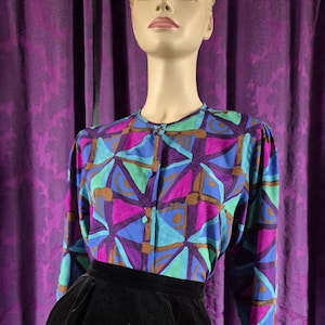 May include: A vintage blouse and skirt set on a mannequin. The blouse features a geometric pattern in shades of blue, purple, green, and brown. The skirt is a high-waisted, black velvet design. The outfit is displayed against a purple curtain backdrop.