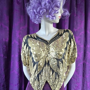 Op de afbeelding: Een mannequin toont een goudkleurige top met pailletten en een vlinderontwerp over een paarse geplooide rok. De top heeft zwarte accenten en korte mouwen. De mannequin heeft een paarse pruik en staat tegen een paarse achtergrond.