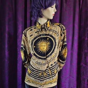Könnte beinhalten: Langärmliges Hemd mit einem Himmelsdesign. Das Hemd hat einen beigefarbenen Hintergrund mit einer großen Sonnengrafik und astrologischen Symbolen in Schwarz und Gold. Das Hemd wird mit einem schwarzen Rock kombiniert.