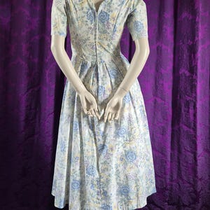 May include: A floral print dress on a mannequin. The dress has a V-neck, short sleeves, and a pleated skirt. The pattern includes blue, yellow, and white flowers on a light background. The dress is made of a light fabric, suitable for warmer weather.