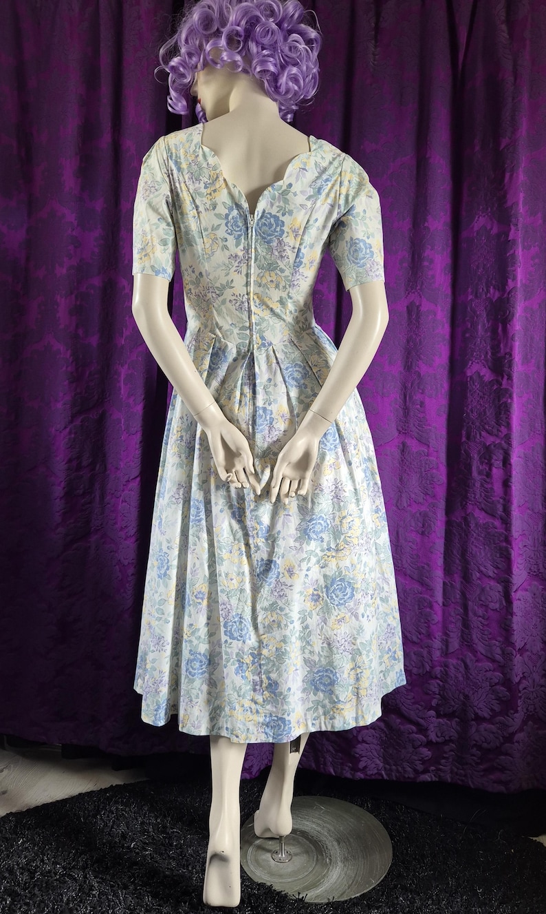 May include: A floral print dress on a mannequin. The dress has a V-neck, short sleeves, and a pleated skirt. The pattern includes blue, yellow, and white flowers on a light background. The dress is made of a light fabric, suitable for warmer weather.