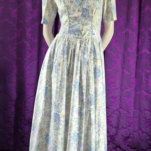 May include: A vintage floral dress with a scoop neckline and short sleeves. The dress has a button-down bodice and a pleated skirt. The fabric is white with blue, yellow, and purple floral patterns. The dress is displayed on a mannequin.