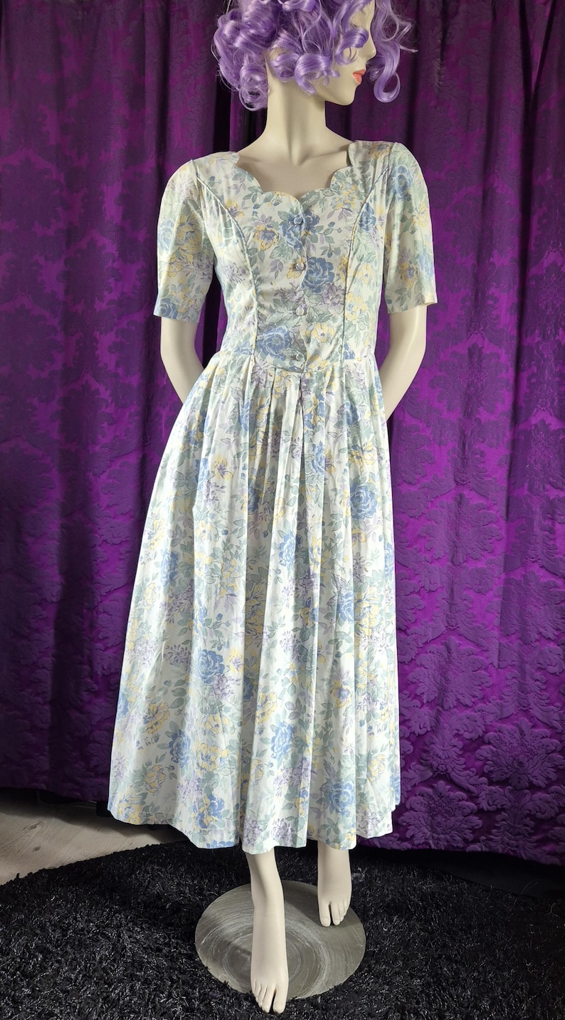May include: A vintage floral dress with a scoop neckline and short sleeves. The dress has a button-down bodice and a pleated skirt. The fabric is white with blue, yellow, and purple floral patterns. The dress is displayed on a mannequin.
