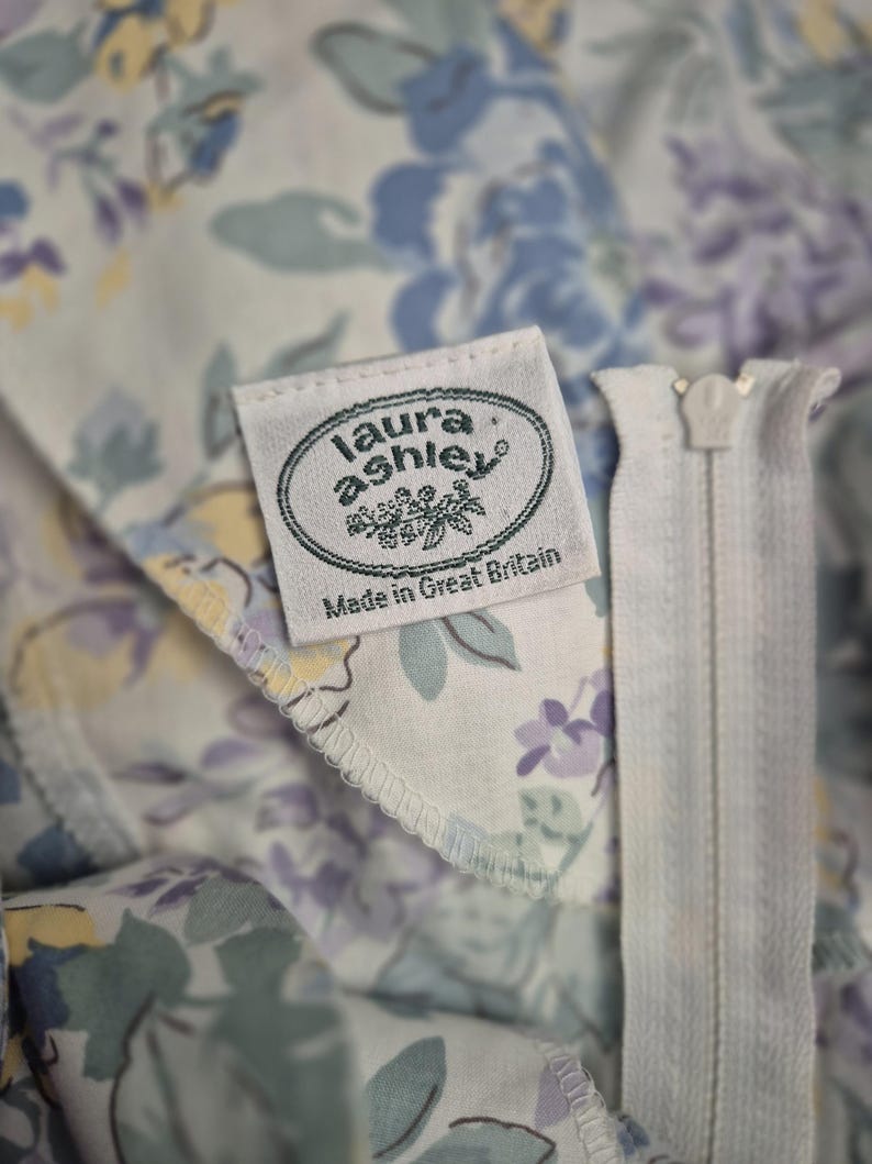 May include: Close-up of a floral fabric with a white zip. The fabric displays a pattern of blue, purple, and yellow flowers on a light background. A label reads "laura ashley Made in Great Britain."