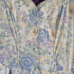 May include: Close-up of a light-coloured floral dress with a button-up front. The dress features a pattern of blue, yellow, and purple flowers with green leaves. The fabric has a soft, slightly textured appearance, and the dress has a scalloped neckline.