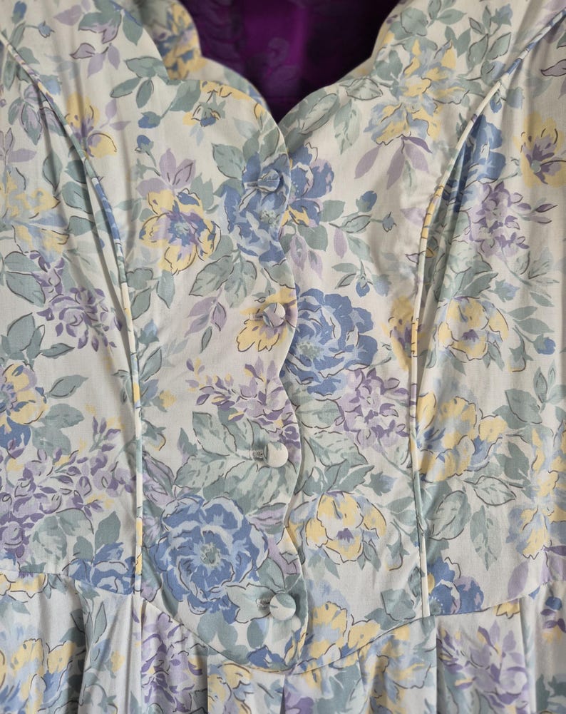 May include: Close-up of a light-coloured floral dress with a button-up front. The dress features a pattern of blue, yellow, and purple flowers with green leaves. The fabric has a soft, slightly textured appearance, and the dress has a scalloped neckline.