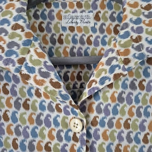 May include: A button-down shirt with a paisley pattern in shades of blue, green, brown, and purple. The shirt has a collar and is hanging on a wooden hanger. The label reads "alexon Liberty Prints."