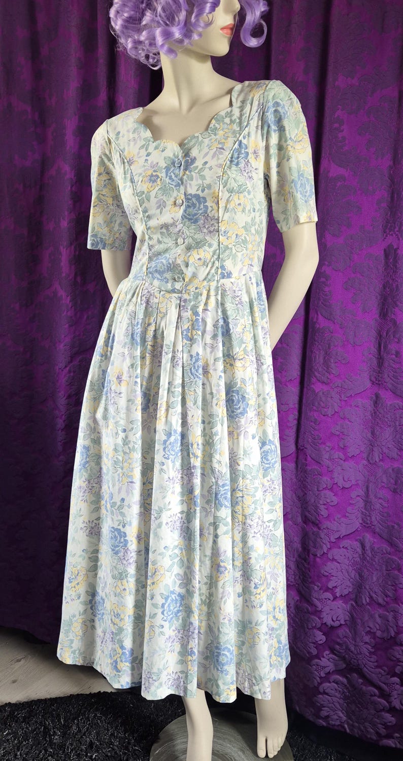 May include: A vintage floral print dress with a scalloped neckline and short sleeves. The dress has a button-down bodice and a pleated skirt. The fabric is white with blue, yellow, and purple floral patterns. The dress is displayed on a mannequin.