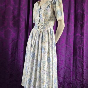 May include: A vintage floral dress in white with blue, yellow, and purple flowers. The dress has short sleeves, a button-up bodice, and a flared skirt. The dress is displayed on a mannequin. The dress is a classic style.