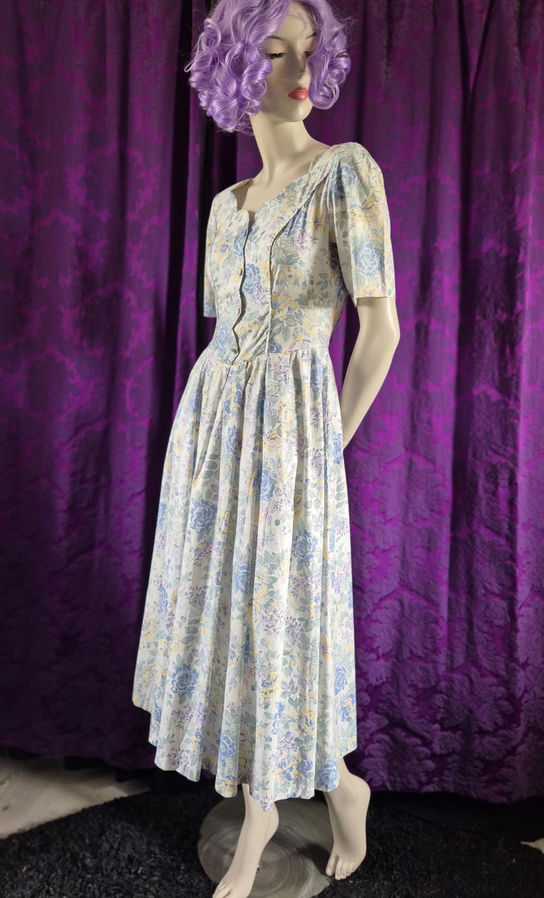 May include: A vintage floral dress in white with blue, yellow, and purple flowers. The dress has short sleeves, a button-up bodice, and a flared skirt. The dress is displayed on a mannequin. The dress is a classic style.