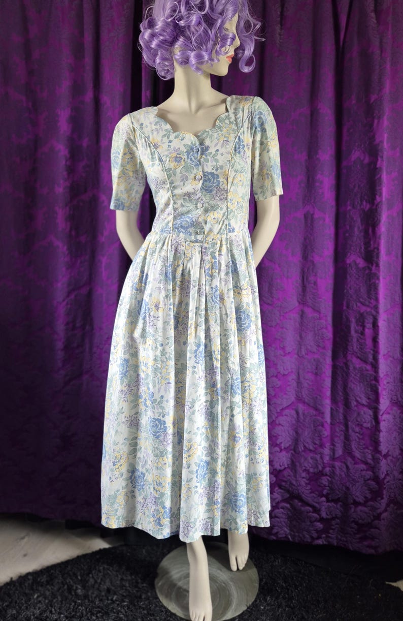 May include: A vintage floral dress with a white base and blue, yellow, and purple flower patterns. The dress has short sleeves, a fitted bodice, and a flowing skirt. It is displayed on a mannequin, with a purple curtain backdrop.