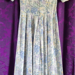May include: A vintage floral dress in pastel shades. The dress has short sleeves, a button-down bodice, and a pleated skirt. The floral pattern features blue, yellow, and purple flowers on a white backdrop. The dress is approximately 130 cm in length.