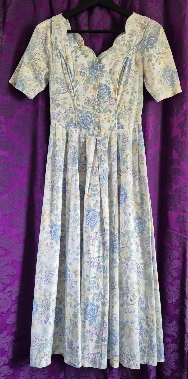 May include: A vintage floral dress in pastel shades. The dress has short sleeves, a button-down bodice, and a pleated skirt. The floral pattern features blue, yellow, and purple flowers on a white backdrop. The dress is approximately 130 cm in length.