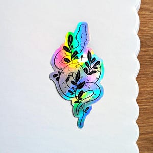 May include: Holographic sticker with a snake design and black leafy branches. The sticker has a rainbow sheen, with colours shifting from blue to yellow and green. The snake is outlined in black.