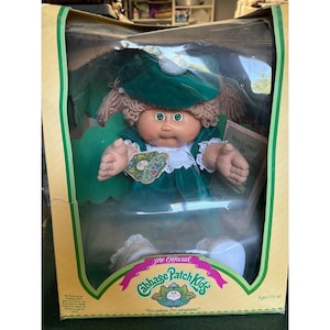 Peut inclure: Une poupée Cabbage Patch Kids avec des cheveux en laine, des yeux verts et un visage rond, portant une robe en velours vert foncé avec un col en dentelle blanche et un chapeau vert assorti. La poupée est dans une boîte avec le texte The Official Cabbage Patch Kids.