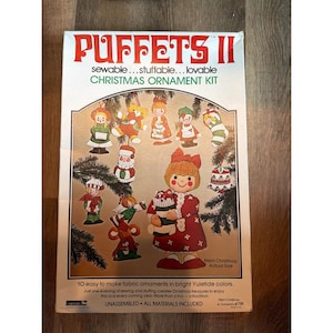 Könnte beinhalten: Eine Vintage-Weihnachtsschmuck-Kit-Box mit dem Titel "PUFFETS II". Die Box zeigt Illustrationen von Stoffornamenten in leuchtenden Farben, darunter Weihnachtsmänner, Elfen und Schneemänner. Der Text lautet "sewable...stuffable...lovable CHRISTMAS ORNAMENT KIT."