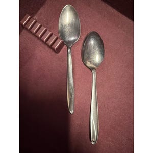 Op de afbeelding: Twee roestvrijstalen lepels met ovale kommen en lange, slanke handvatten. Eén lepel heeft een decoratieve werveling op het handvat. Een donkere chocoladereep is ook zichtbaar in de afbeelding. De items staan op een bordeauxrode ondergrond.