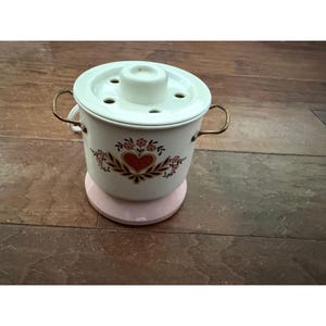 Op de afbeelding: Een vintage witte en roze elektrische voedselstomer. De stomer heeft een wit deksel met ventilatiegaten en een centrale knop. De behuizing is versierd met een hart- en bloemenontwerp. Het heeft goudkleurige handvatten.