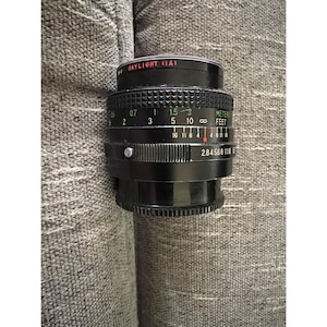 May include: A black camera lens with the text "SKYLIGHT (IA)". The lens has a ridged focus ring with distance markings in meters and feet. Aperture settings are also visible. The lens is set against a neutral background.