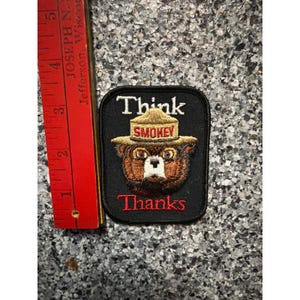 May include: A rectangular black embroidered patch featuring a brown bear wearing a tan ranger hat with the text 'SMOKEY' in red, 'Think' in white, and 'Thanks' in red. A red ruler is placed next to the patch, showing a height of 3 inches.