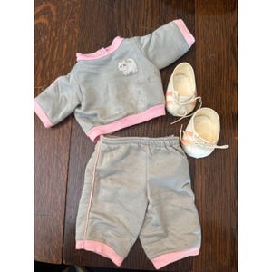 Könnte beinhalten: Ein graues Puppen-Outfit mit rosa Besatz, bestehend aus einem Sweatshirt mit Katzenapplikation, Shorts und einem Paar weißer Schuhe mit rosa Streifen. Das Outfit ist für eine Puppe bestimmt.
