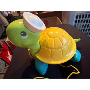 May include: A green and yellow toy turtle with a white sailor hat. The turtle has blue feet and a yellow pull string. The turtle's shell is yellow and has a textured design.