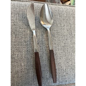 May include: A stainless steel butter knife and spoon set with dark brown wooden handles. The knife has a rounded blade, and the spoon has a deep bowl. The utensils are placed on a gray textured surface.