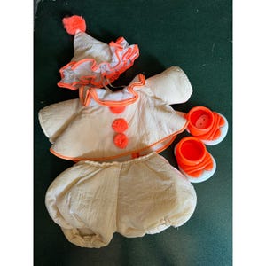 May include: A cream-colored clown costume set consisting of a cone-shaped hat with a bright orange pom-pom, a ruffled collar, a long-sleeved top with two orange pom-poms, matching bloomers, and a pair of bright orange plastic doll shoes.